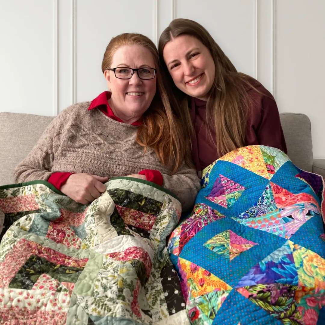 Cheryl and Marsali sitting together on a couch, each holding a quilt