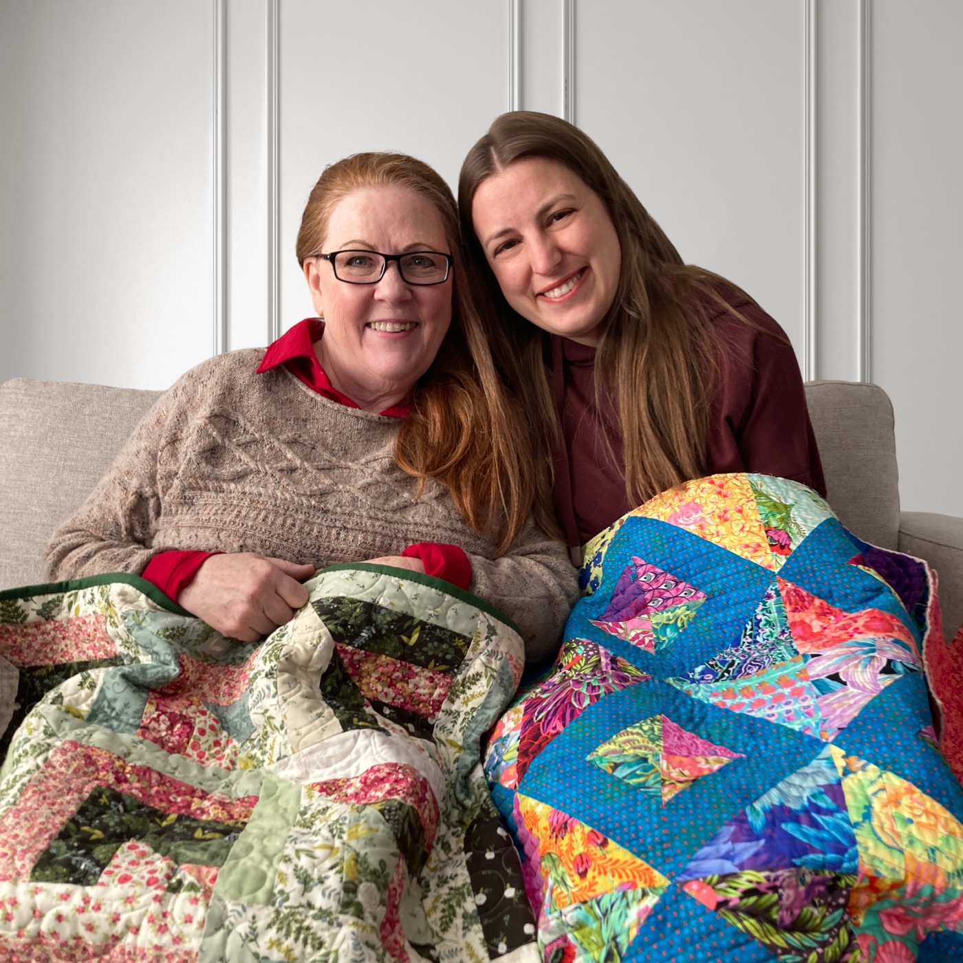 Cheryl and Marsali sitting together on a couch, each holding a quilt